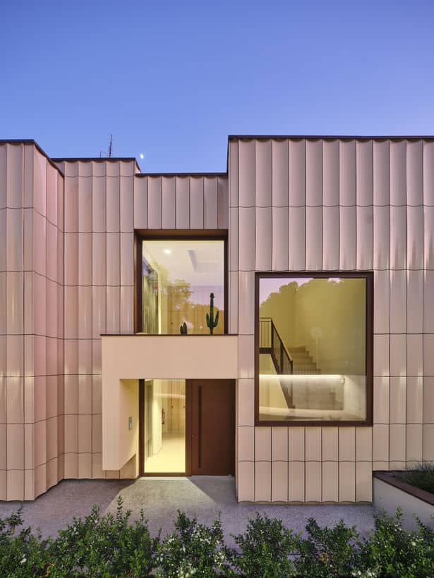 Modern residential fa&ccedil;ade with vertical beige ceramic tiles and large windows revealing a warmly lit interior at dusk.