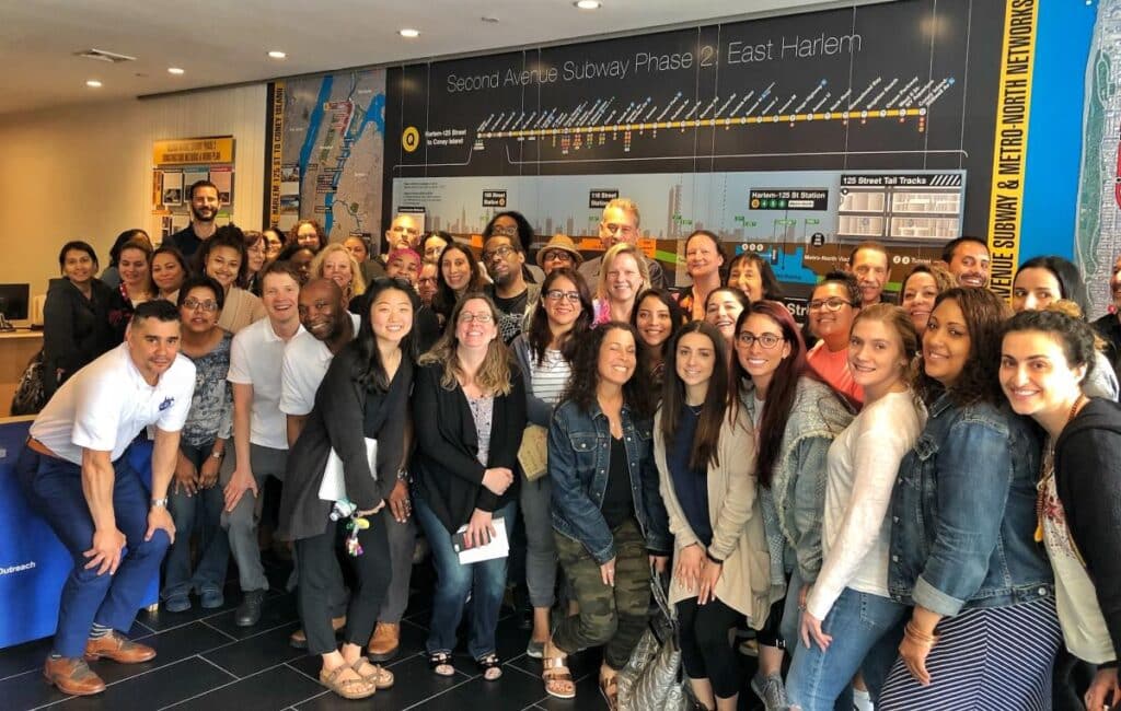 A diverse group of people &mdash; staff, community members, and stakeholders &mdash; smiling together in front of a large display showing the Phase 2 map of the Second Avenue Subway expansion into East Harlem.