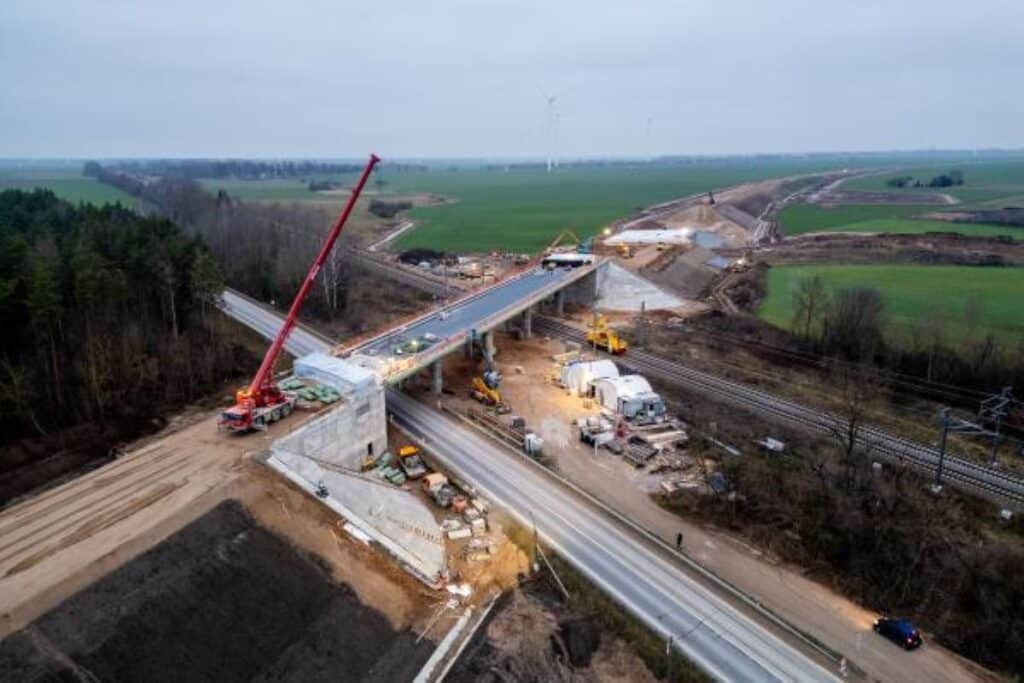 An aerial view of a new bridge under construction as part of the Rail Baltica project, featuring a giant red crane installing segments over a road and railway line, surrounded by green farmland and bare trees &mdash; hinting at early spring or late winter.