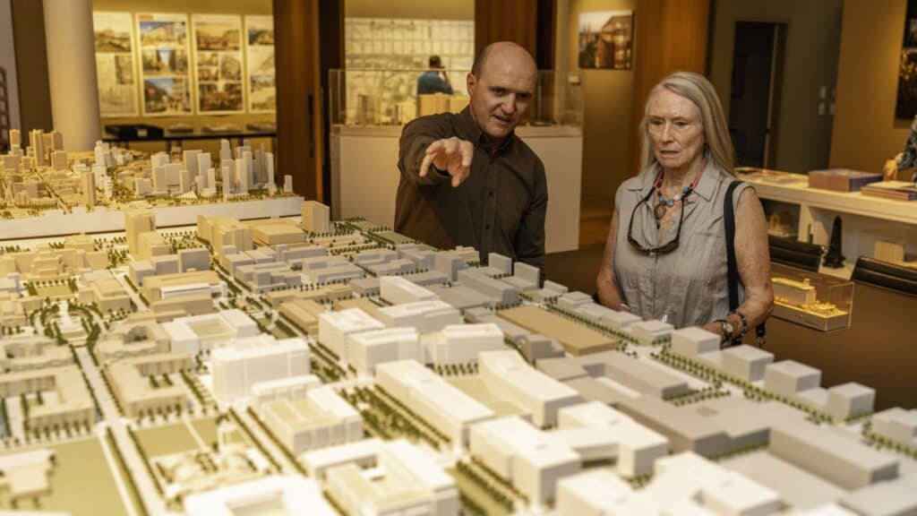 A man points to a detailed city model while explaining it to a woman during a Doors Open Denver event &mdash; an intimate moment of learning and discovery in a warmly lit gallery.