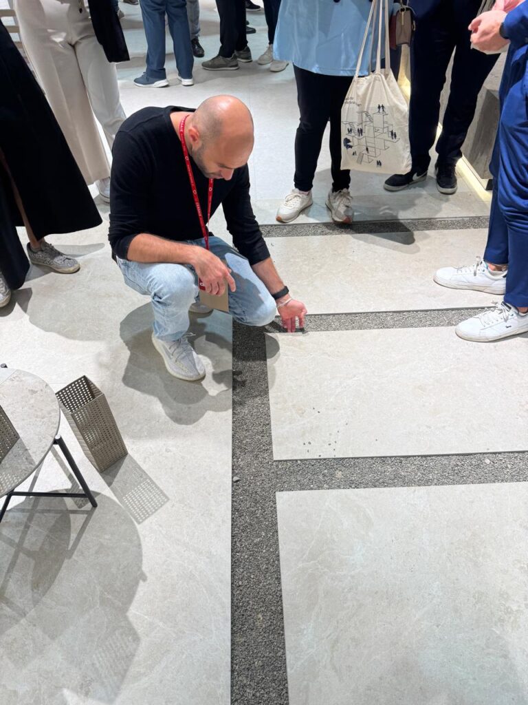 رجل يركع على الأرض في معرض Cersaie 2025 ليتفحّص عن قرب تصميم وأسطح بلاطات السيراميك، بينما يحيط به زوار آخرون يتابعون العرض بانتباه.