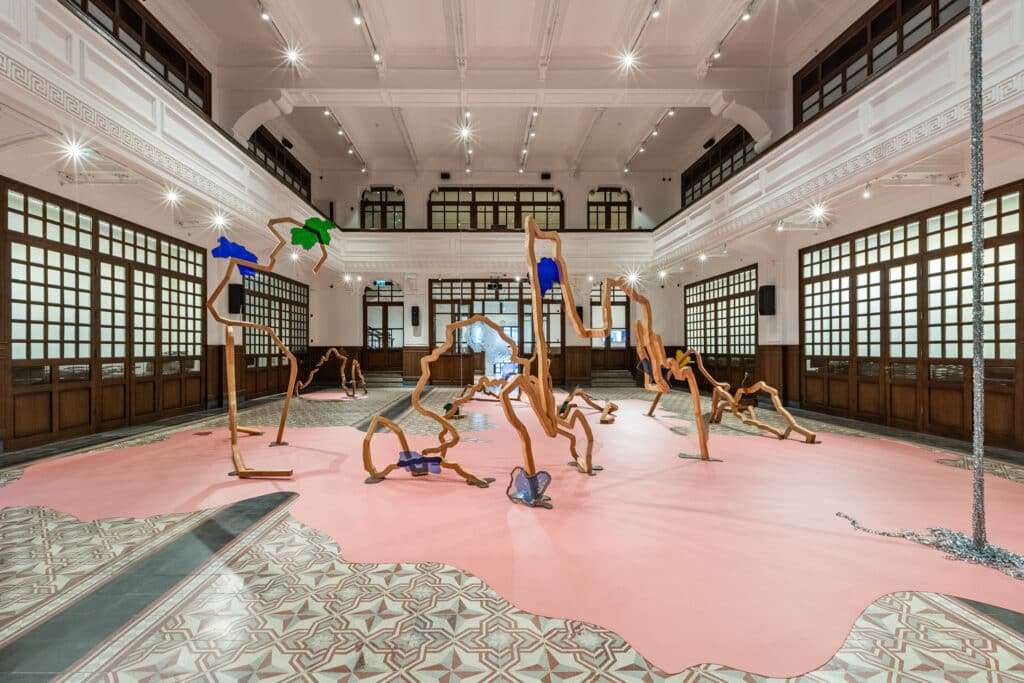 A large, dreamlike sculpture installation with wooden limbs and colorful accents sprawls across a pink floor inside a grand historic hall with ornate ceilings and patterned tiles.