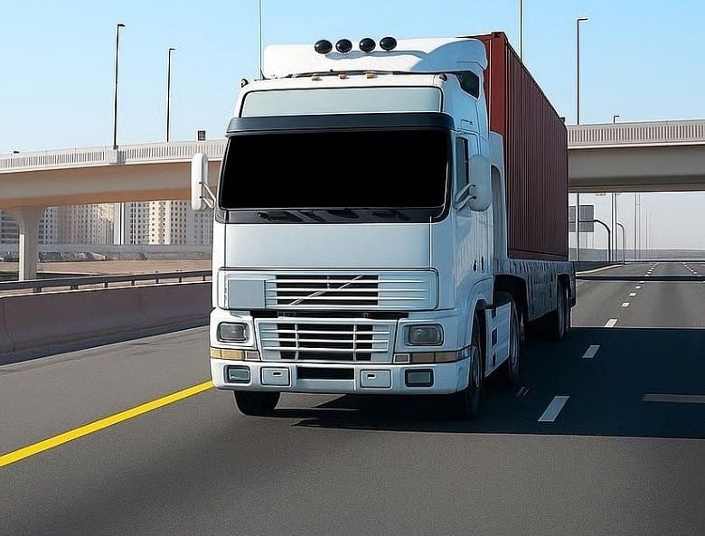 A white autonomous semi-truck hauling a red shipping container cruises down a sunlit highway in Dubai, with modern overpasses and urban skyline in the background.