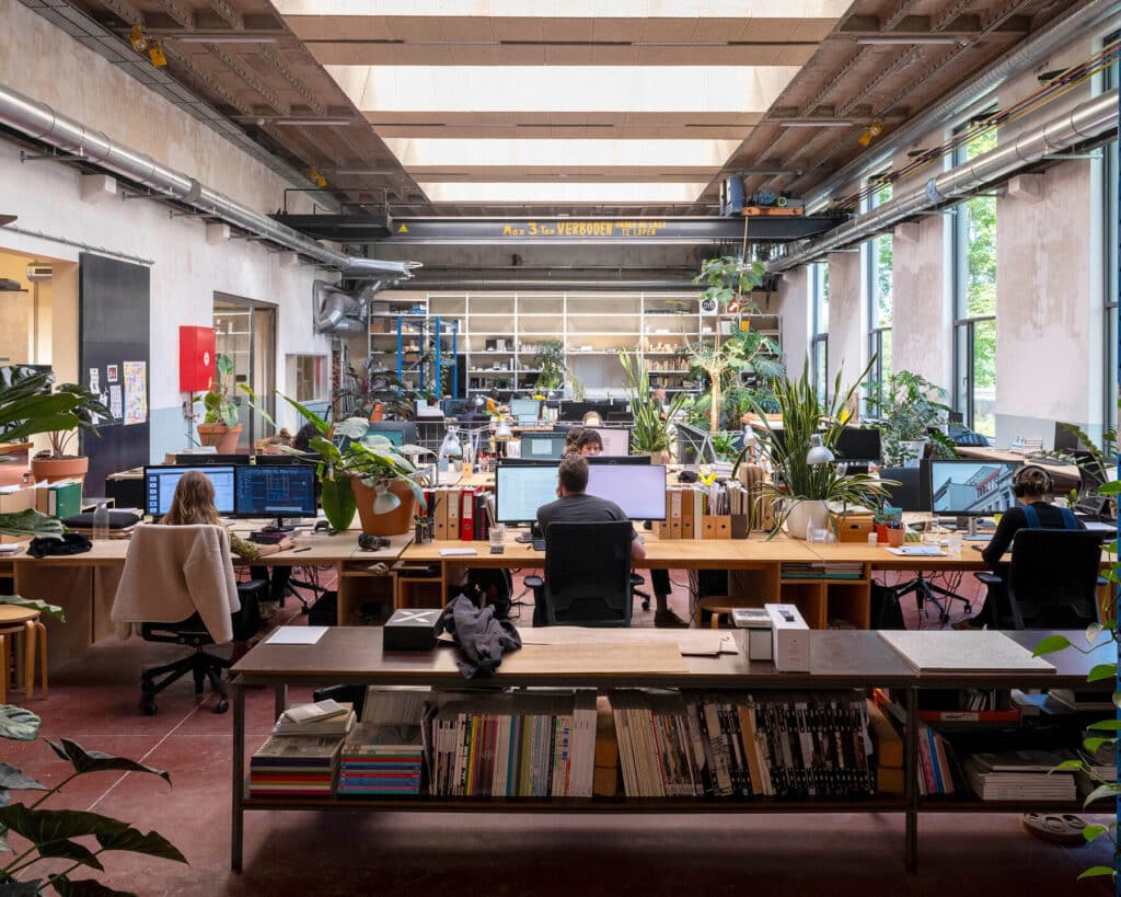 A vibrant architectural studio with employees working at wooden desks surrounded by large indoor plants. Exposed industrial elements like metal pipes and a vintage overhead crane marked &ldquo;Max 3 Ton VERBODEN&rdquo; reveal the building&rsquo;s former industrial life. Natural light streams in through tall windows, while bookshelves and storage units line the space, blending functionality with greenery.