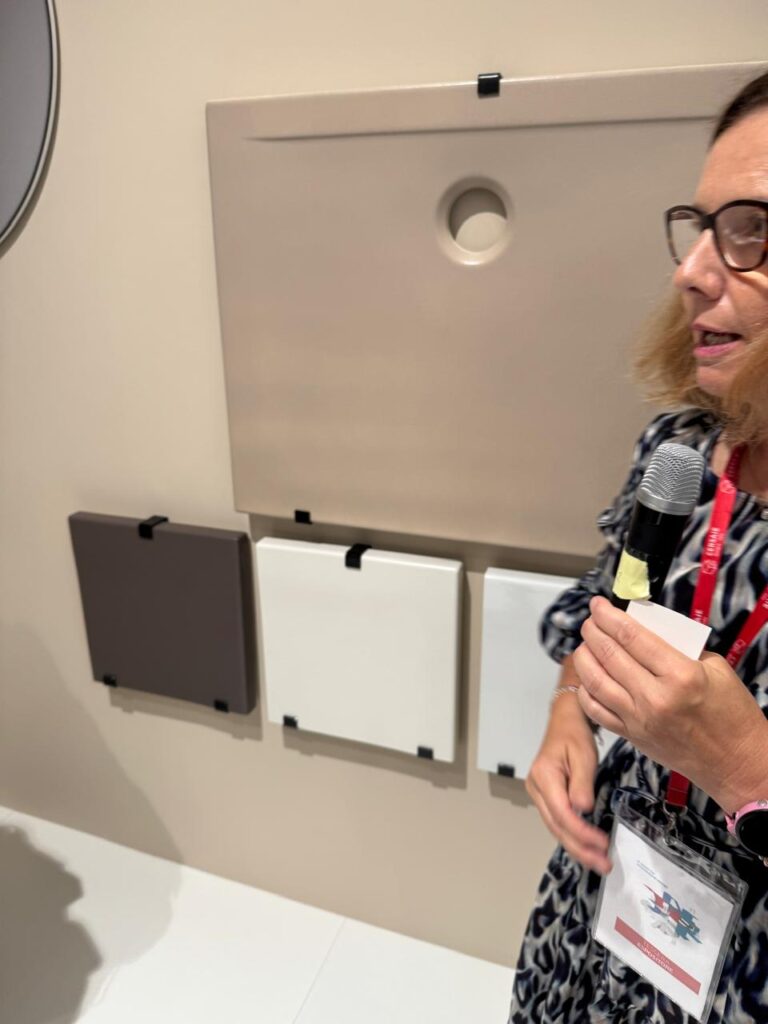 A woman with glasses and a lanyard speaks into a microphone while presenting sleek, modern wall-mounted fixtures at Cersaie 2025. Behind her, minimalist panels in soft tones showcase design details &mdash; a quiet moment of expert storytelling in the heart of the fair.