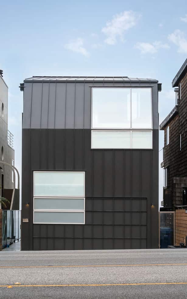 A modern, box-like house with dark gray metal cladding and geometrically arranged glass windows, standing on a coastal street between neighboring homes.
