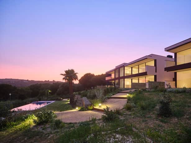 Modern residential buildings on a hillside with an infinity pool, illuminated at dusk under a pink and purple sky.