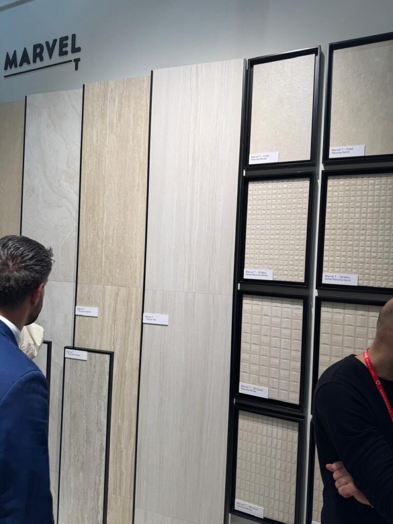 Two visitors examine a display of “Marvel T” porcelain panels at Cersaie 2025, featuring a curated mix of wood-look, stone-effect, and 3D textured tiles in soft whites and beiges.