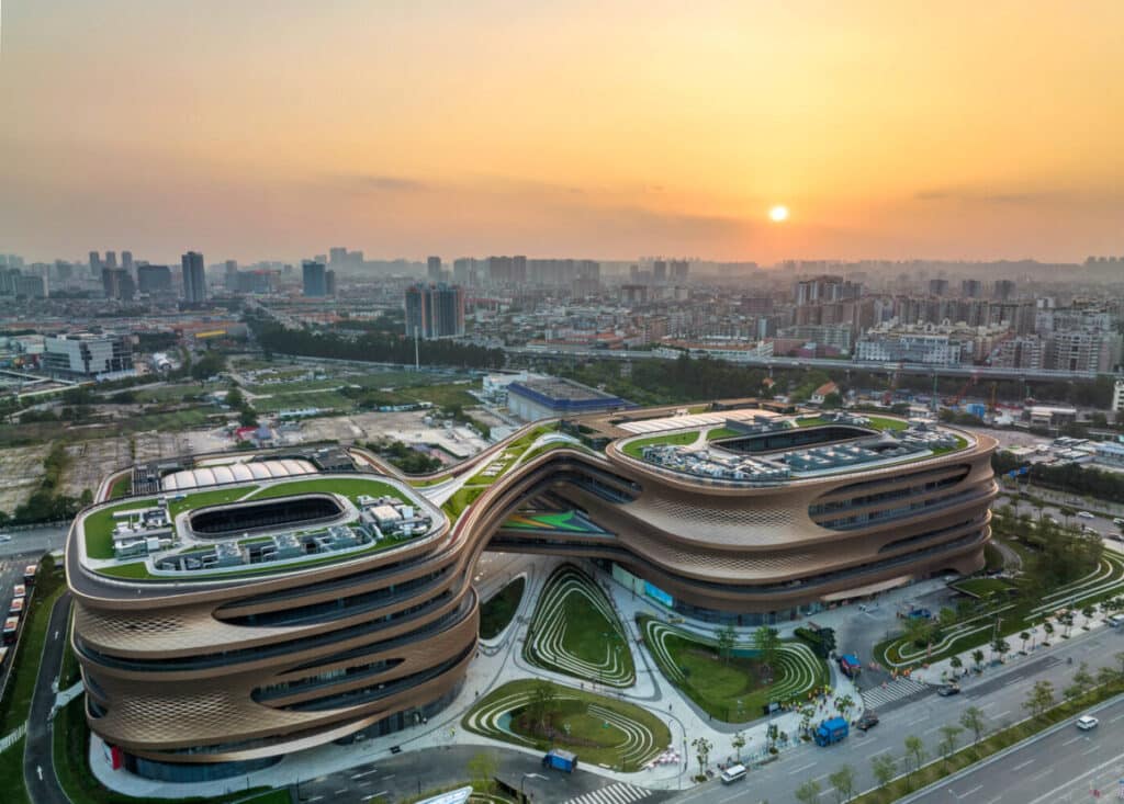 An aerial view of a futuristic architectural complex with flowing, ribbon-like forms and green rooftops, set against a golden sunset. The building features interconnected curved structures surrounded by landscaped gardens and urban infrastructure.