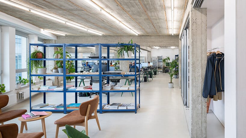 A modern, open-plan architectural office with blue metal shelving units filled with books and documents, interspersed with indoor plants. The exposed concrete ceiling features linear lighting, while white walls and polished floors create a clean, airy atmosphere. Wooden chairs and minimalist furniture add warmth to the functional space.