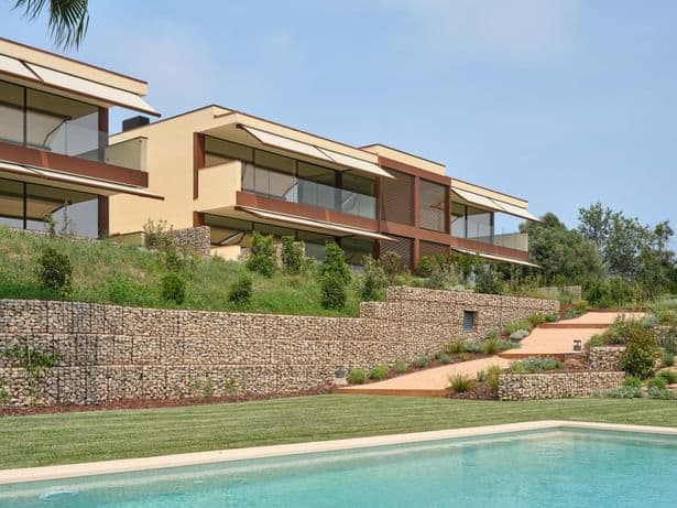 Modern residential units built on a hillside with an infinity pool in the foreground, surrounded by stone retaining walls and natural landscaping.