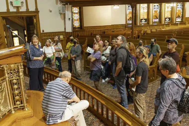 A group of visitors gathered inside a historic church with wooden pews and stained-glass windows, listening intently to a guide during a Doors Open Denver tour.