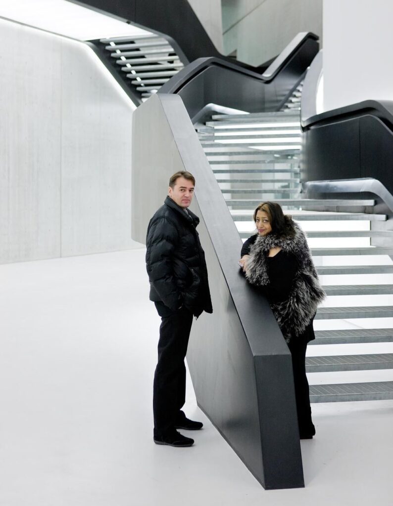 A man and a woman stand in a minimalist, futuristic architectural space with sculptural staircases and sleek black and white surfaces. The clean lines and dramatic geometry highlight the bold design of the interior.