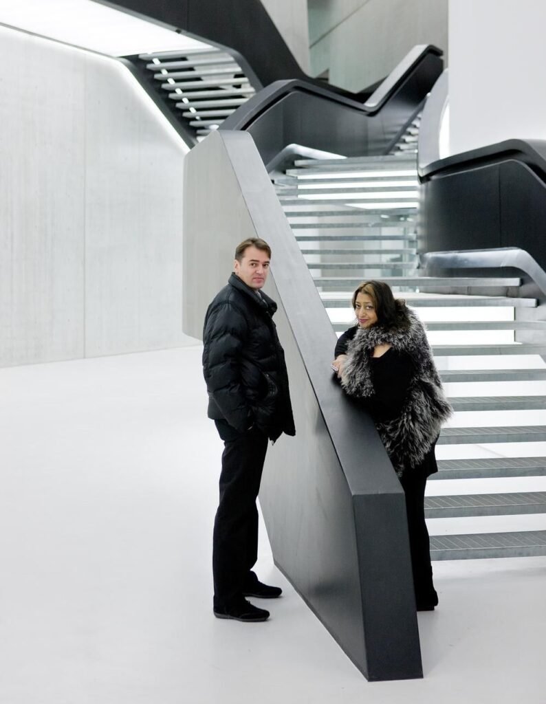 رجل وامرأة يقفان داخل مساحة معمارية حديثة ذات تصميم مينيماлист، مع سلالم معلقة وحوائط بألوان سوداء وبيضاء نظيفة. الخطوط المستقيمة والهندسة الجريئة تعكس التصميم المبتكر للداخل.