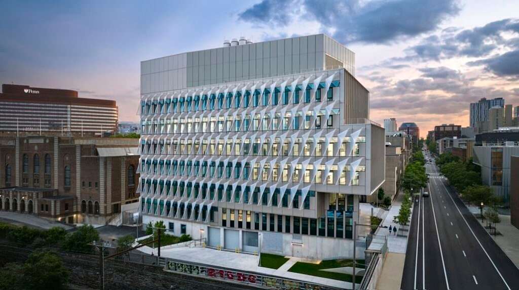 The Vagelos Laboratory for Energy at UPenn at sunset, featuring its distinctive facade with sculptural white sunshades, glowing interior windows, and an empty street stretching into the city under a dramatic cloudy sky.