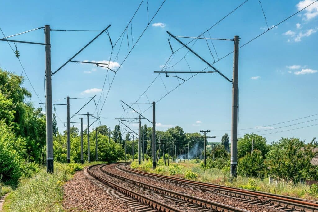 A curving electrified railway track stretching into the distance, flanked by lush green trees and a clear blue sky &mdash; with evenly spaced power poles lining the route, symbolizing harmony between modern infrastructure and natural beauty.