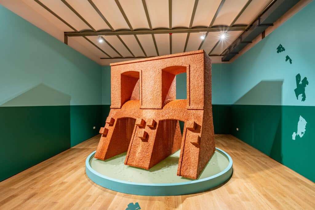 A bold, terracotta-colored architectural sculpture with arches and textured surfaces stands on a circular platform in a gallery with mint-green walls and wooden floors &mdash; evoking ancient ruins reimagined through modern art.