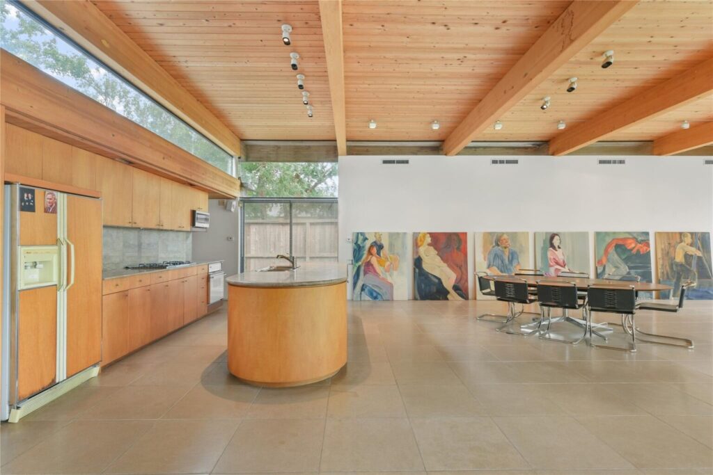 مطبخ ومجالس مفتوحة بسقف خشبي وطاولة مستديرة في المنتصف، مع لوحات فنية كبيرة على الحائط ونوافذ طويلة تسمح بدخول الضوء الطبيعي.