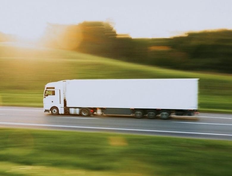 A white semi-truck speeds down a highway at golden hour, with blurred green fields and warm sunlight painting the scene &mdash; capturing the motion and efficiency of modern logistics.