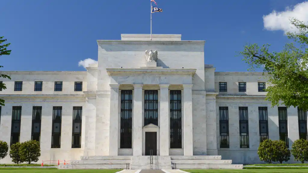 The U.S. Federal Reserve Building bathed in sunlight, showcasing its majestic marble facade, towering columns, and the iconic eagle statue above the entrance. The American flag and Fed flag flutter atop, framed by lush green trees under a bright blue sky.