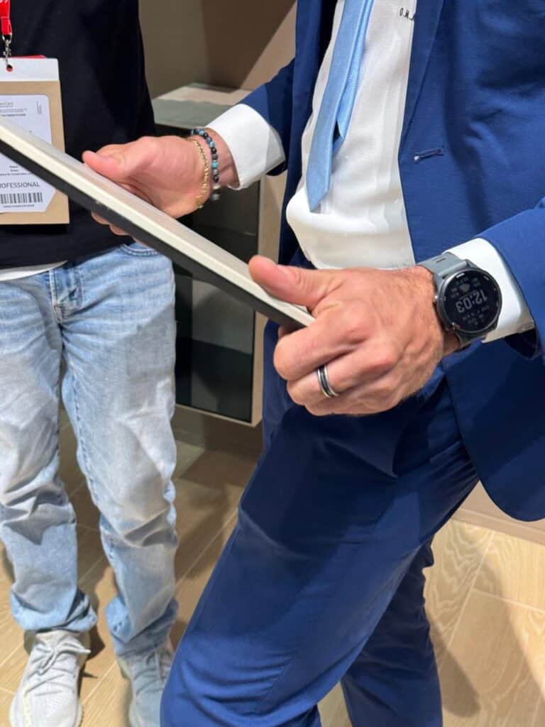 رجل في بدلة زرقاء يحمل قطعة بلاطة سيراميك بين يديه، يُظهر تفاصيل الحافة والسمك أثناء مناقشتها مع زميل، في جو احترافي ومُلهم من معرض Cersaie 2025.