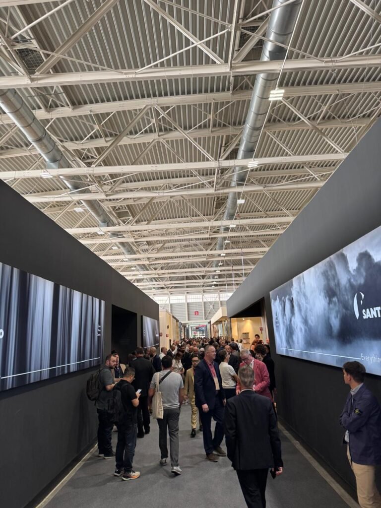 A bustling corridor at Cersaie 2025, packed with attendees walking between booths under an industrial-style steel ceiling, with large digital screens displaying brand visuals like “SANT” on the walls.