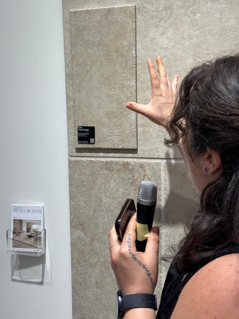 A woman gently touches a large porcelain tile on display at Cersaie 2025, holding a microphone in one hand &mdash; likely explaining its texture, finish, or technology to an audience. A small label reads &ldquo;Safe Technology,&rdquo; hinting at innovation beneath the surface.