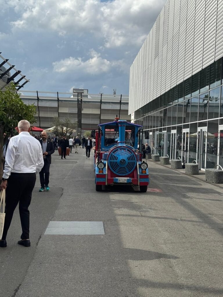 A bright blue and red miniature train rolls down the outdoor walkway of Cersaie 2025, passing by attendees strolling under umbrellas and trees, with the modern glass-and-steel exhibition hall in the background.
