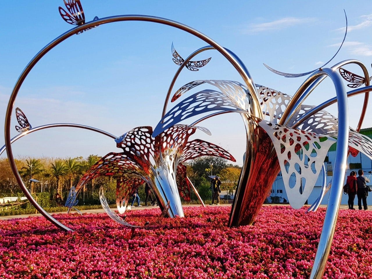 Stainless steel sculpture 'Flowers and Butterflies Dance' reflecting sunlight and surrounding flowers in Taichung, Taiwan