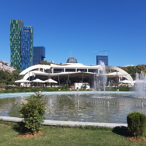 Urban park in Tirana with a central fountain, distinctive modern building, and contemporary towers under a clear sky.