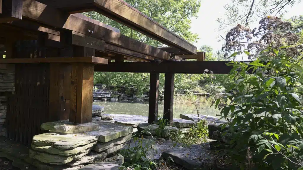 A rustic wooden pavilion with thick beams and stone foundations overlooks a tranquil lily pond, framed by lush greenery and native plants under dappled sunlight.