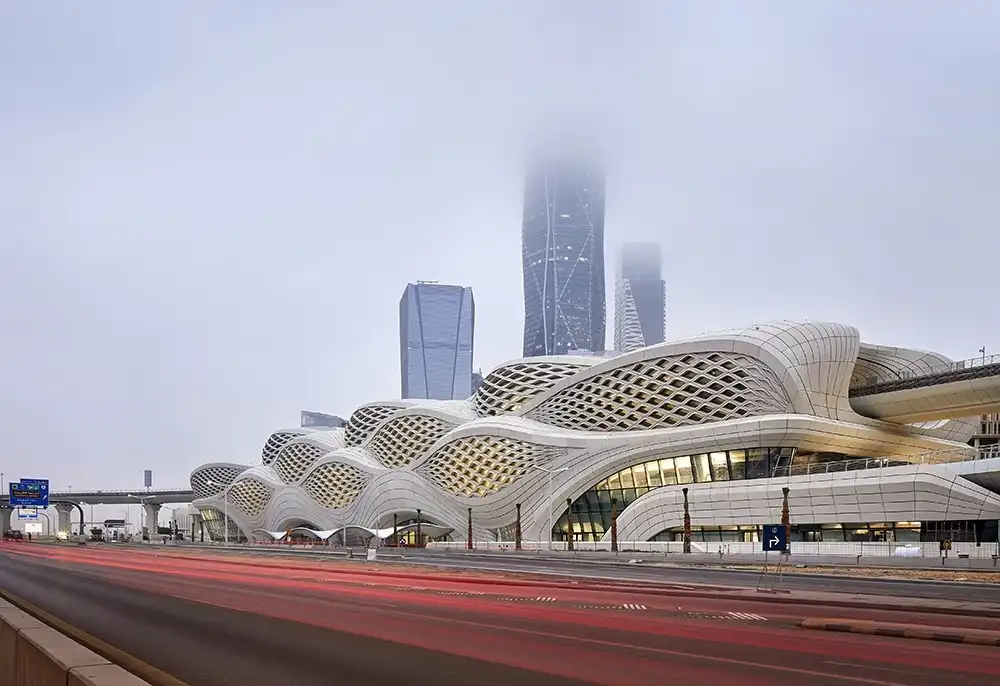 King Abdullah Financial District Metro Station in Riyadh, featuring its iconic wavy concrete lattice facade, with skyscrapers rising behind it through the mist. Red light trails from traffic blur across the foreground, capturing the rhythm of city life.