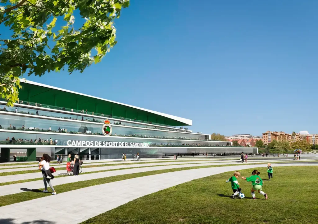 A sunny day at the renovated El Sardinero Stadium, where kids play soccer on the grass in front of the modern glass-and-green building, while people stroll along the wide steps and walkways around it.