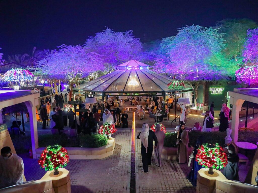 مشهد ليلي مُذهل من فعاليات موسم الرياض 6، يظهر فيه مبنى زجاجي مُضاء بقبة مُزخرفة، وشجر مُضيء بألوان زاهية،