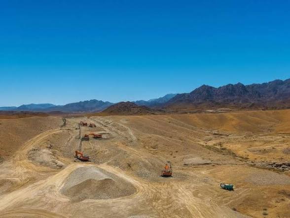 مشهد جوي لآلات ثقيلة تعمل في موقع بناء سكة حديد صحراوية، مع ظهور جبال الحجر الوعرة في الخلفية تحت سماء زرقاء صافية.