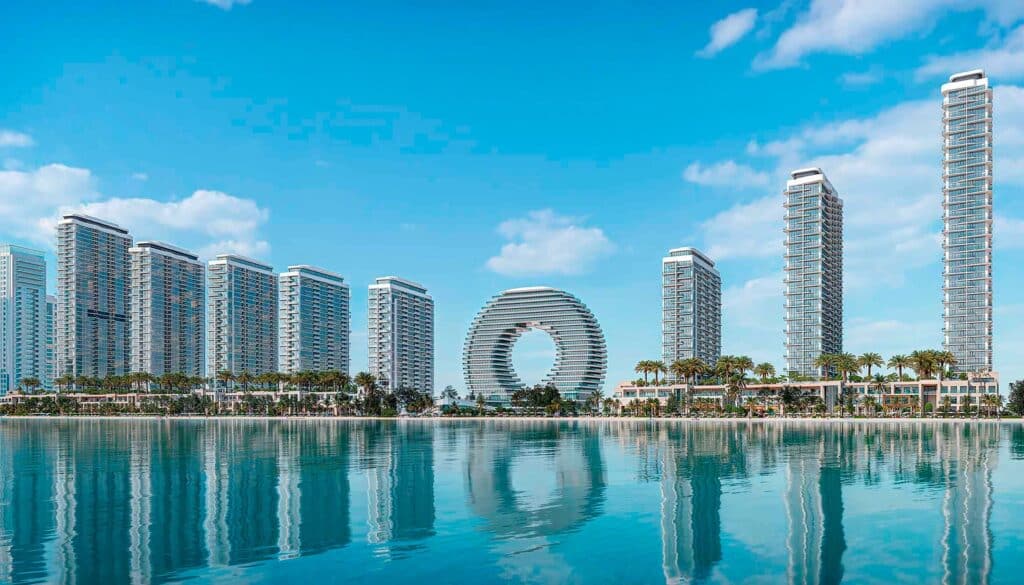 مشهد بانورامي لمدينة ساحلية حديثة في أم القيوين، يظهر أبراجًا سكنية شاهقة ومميزة بتصميمها المعماري العصري، تطل على مياه هادئة تعكس صورتها، مع شاطئ رملي وأشجار نخيل تحت سماء زرقاء صافية.