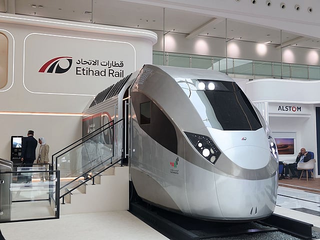 A sleek, futuristic prototype of the Etihad Rail passenger train on display at an official event, with the project&rsquo;s logo visible in the background and visitors observing the design.