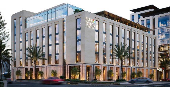 A modern hotel building by Four Points by Sheraton in North Riyadh, captured at dusk with a glass facade, warm interior lighting, palm trees and small shrubs in front, and cars passing by on the street.