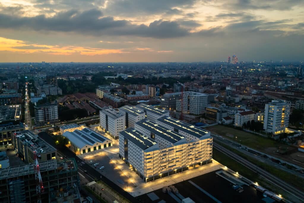 An aerial view of Milan&rsquo;s Olympic Athletes&rsquo; Village at sunset, showcasing modern illuminated buildings alongside historic structures, with the sprawling cityscape and warm-hued sky in the background.