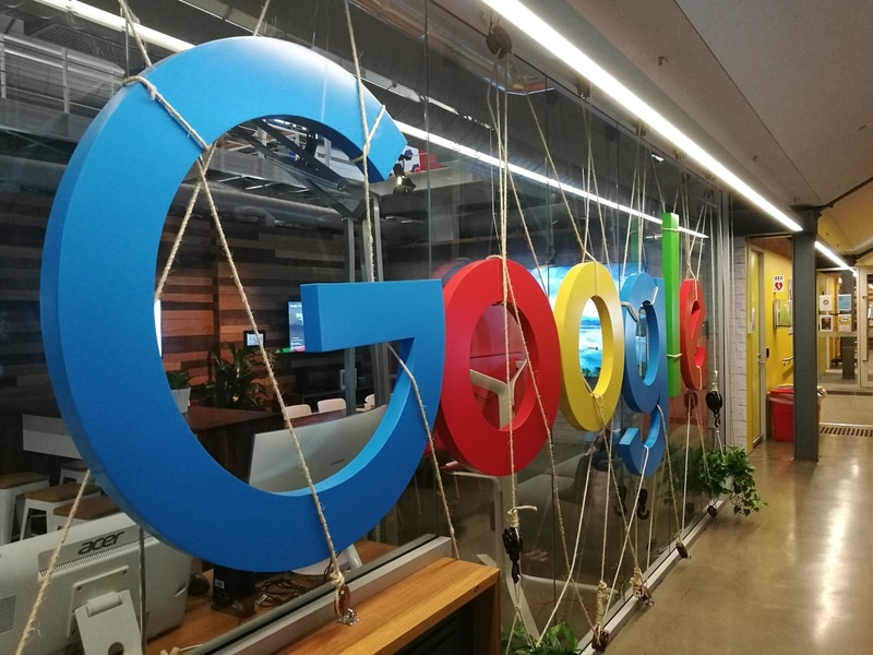A large, colorful Google logo suspended by ropes inside a modern office hallway, with glass walls and plants visible in the background.
