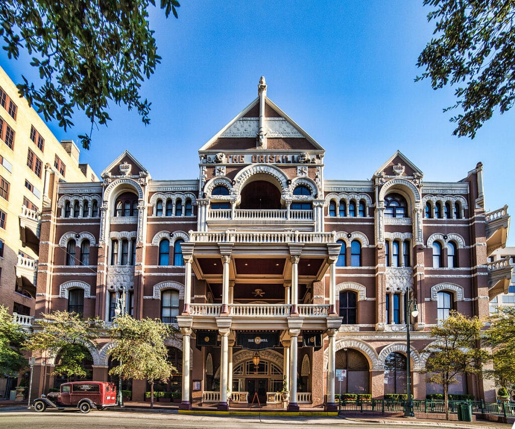 The historic Driskill Hotel in Austin, a red-brick building with classical columns and a vintage car parked out front under a blue sky.