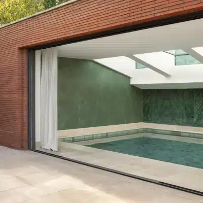 An indoor pool with green walls and skylights, opening to a garden through large glass doors.