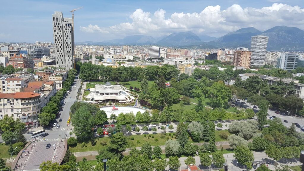 Aerial view of Tirana: a green park nestled between city buildings and distant mountains under a bright sky.