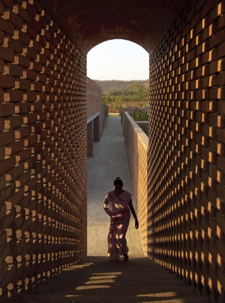 امرأة في ساري وردي تدخل من تحت قوس طوب أحمر مثقب، مع ظلال هندسية على الأرض تحت ضوء الشمس الدافئ.