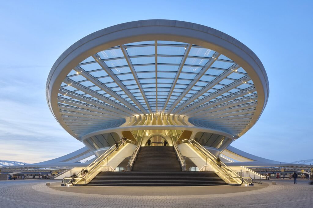Grand glass canopy entrance of Mons station, with lit escalators and stairs.