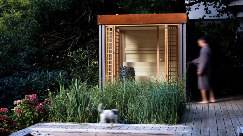ساونا خشبية صغيرة في حديقة، مع باب مفتوح وشخص يمشي بجانبها وكلب صغير يمر أمامها.