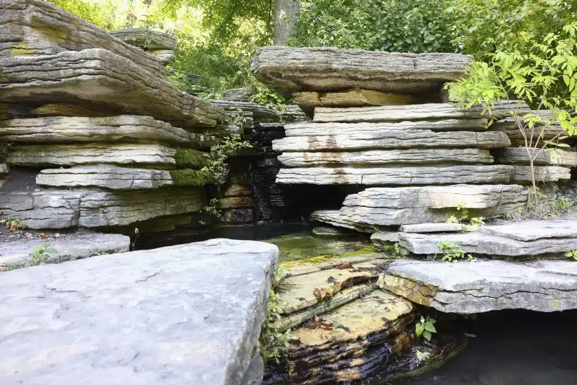 Layered limestone rocks form a natural-looking waterfall and pool, with moss and greenery peeking through the crevices under soft sunlight.