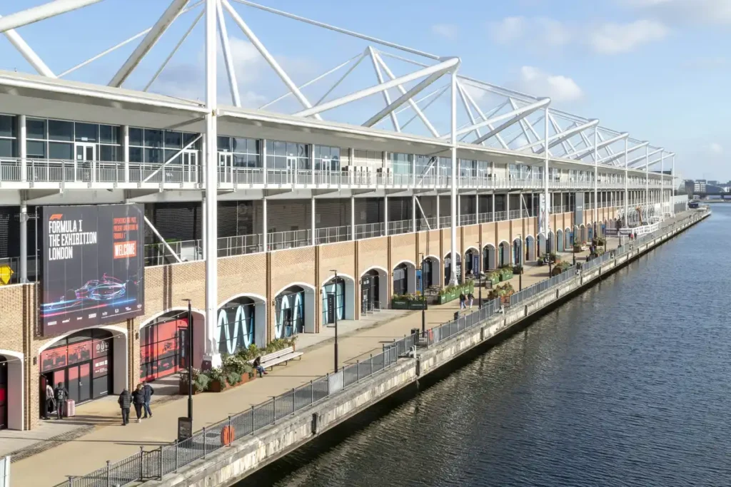 A sunny day at Immerse LDN, where the old car park beneath ExCeL has been reborn as a lively riverside venue. People stroll past arched entrances and modern glass facades, with a giant Formula 1 exhibition banner adding a splash of energy to the brick-lined walkway along the Thames.