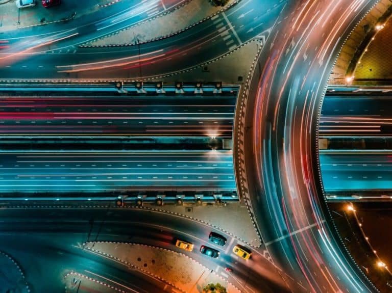 An aerial night shot of a highway interchange, showing streaks of light from moving vehicles across bridges and ramps — blue and red trails painting the rhythm of a city that never sleeps.