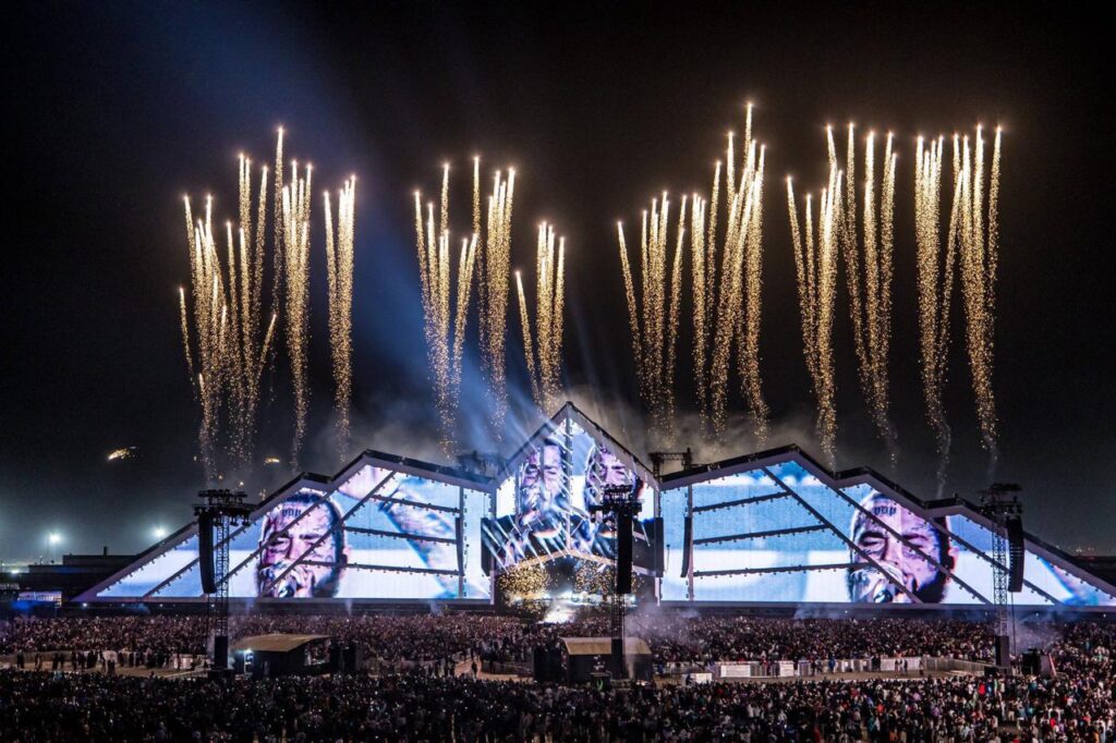 مشهد ليلي مذهل لحفل ضخم في موسم الرياض 6، يظهر فيه مسرح عملاق على شكل هرم مُضاء بشاشات ضخمة تعرض وجه فنان